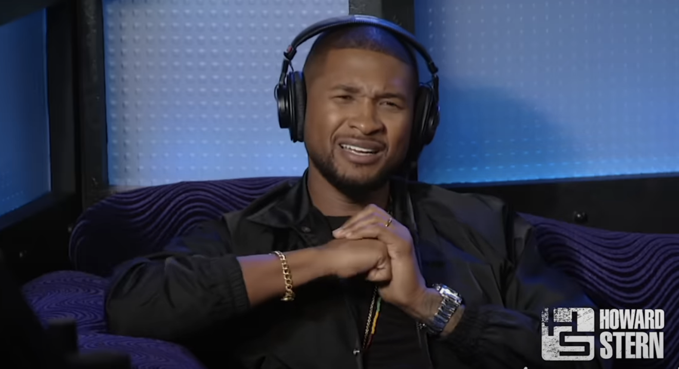 Usher wearing headphones, sitting, smiling with hands clasped