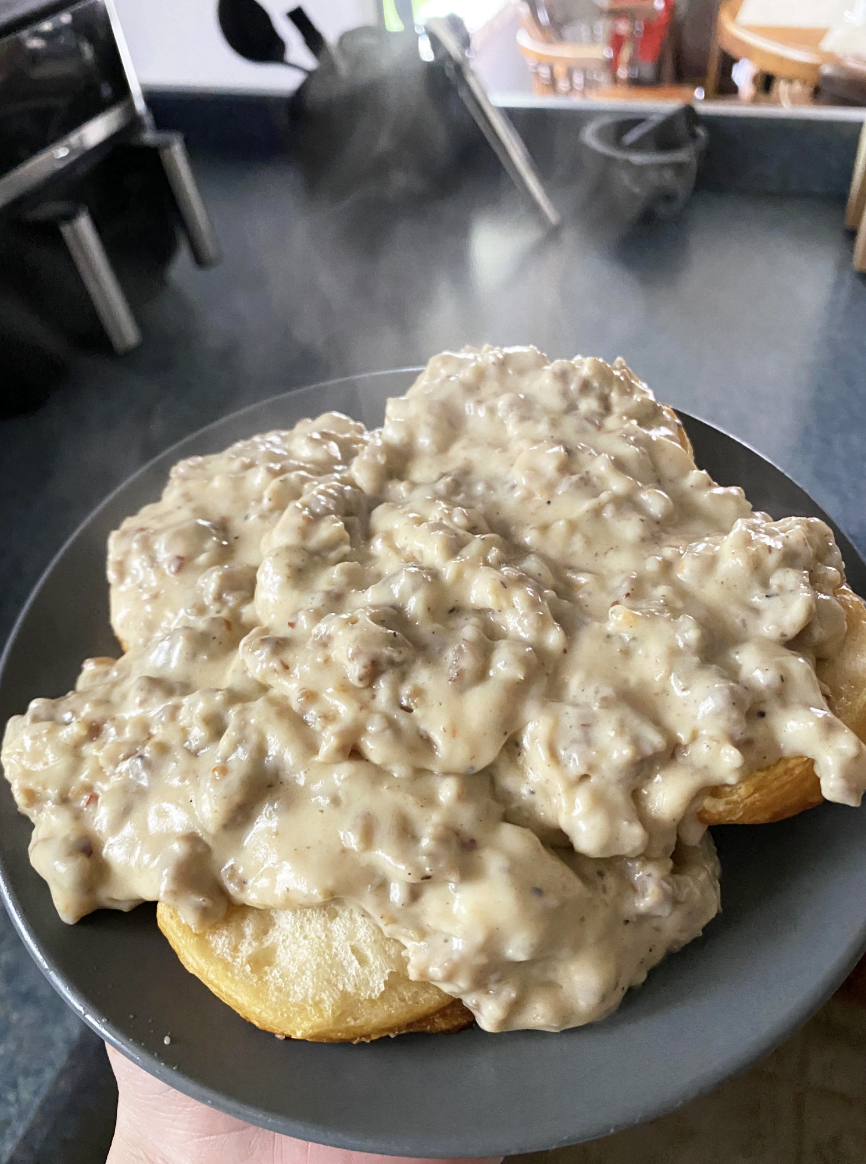 A person holds a plate of biscuits smothered in creamy sausage gravy, steam rising, kitchen background