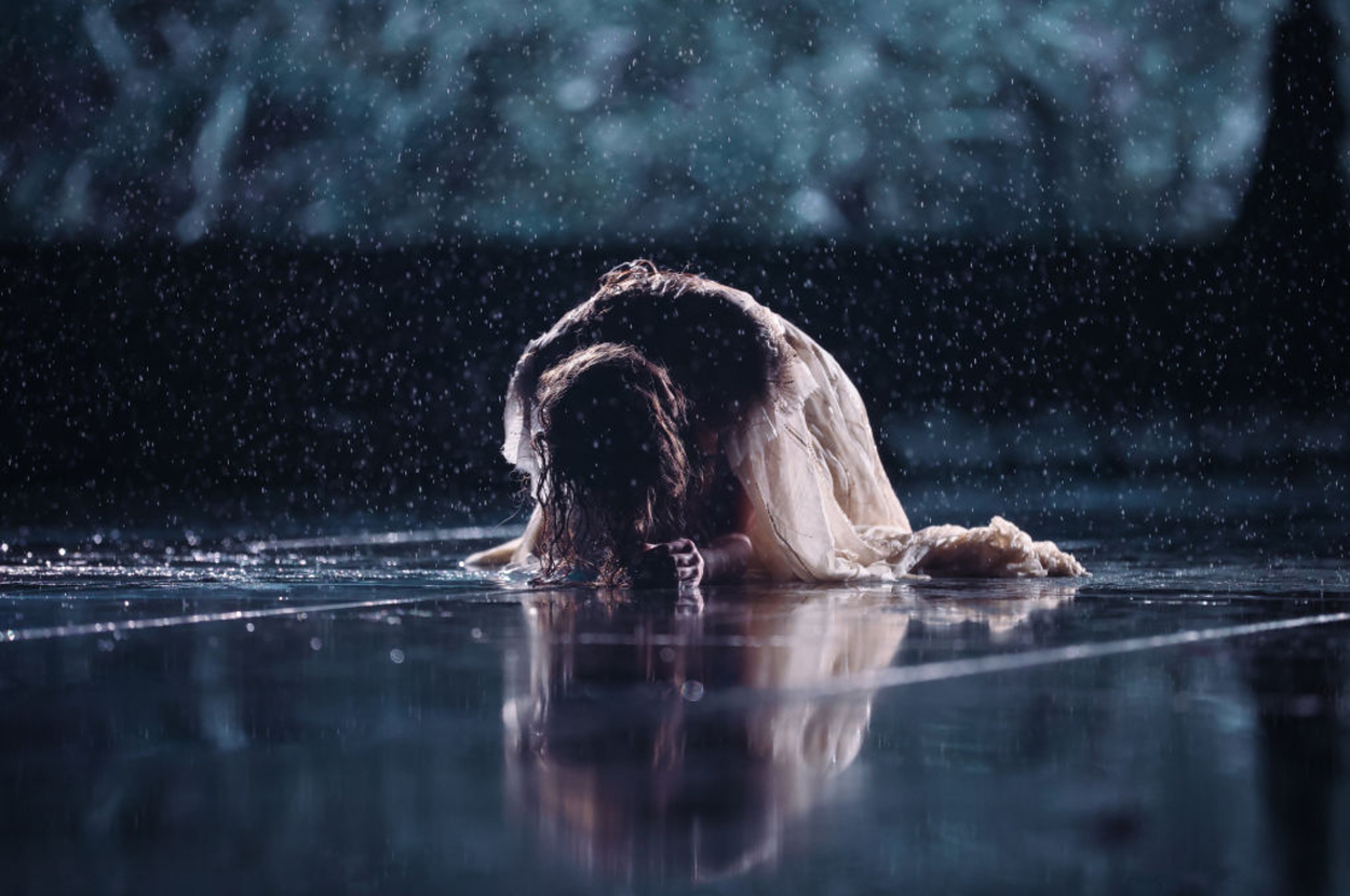Taylor Swift in a flowing dress sitting on wet ground with head bowed, surrounded by raindrops
