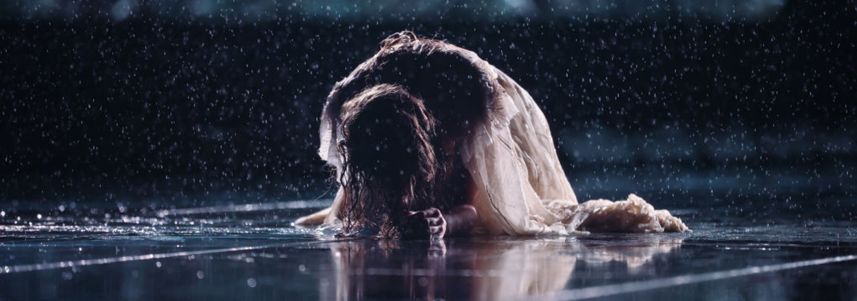 Taylor Swift in a flowing dress sitting on wet ground with head bowed, surrounded by raindrops