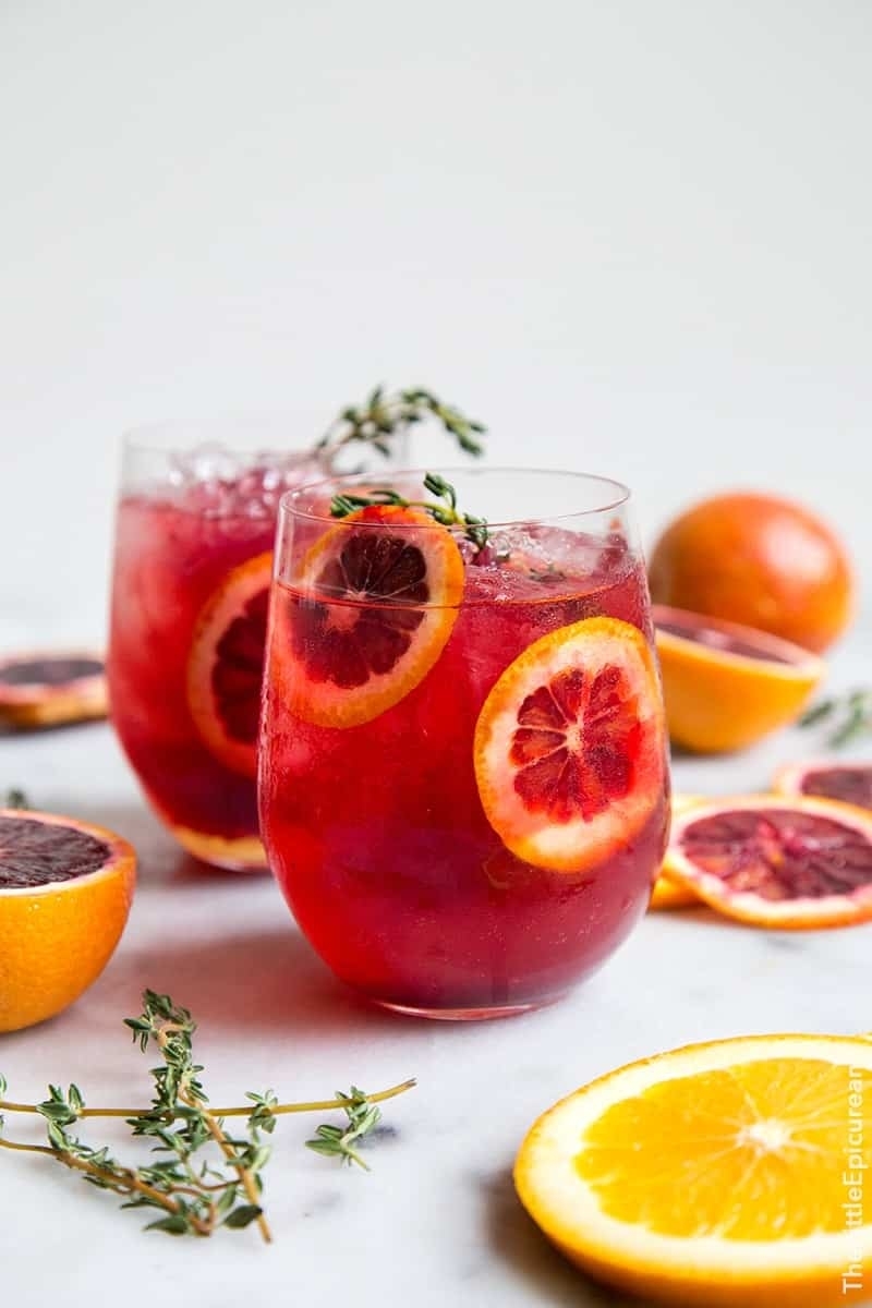 Two glasses with a red beverage and garnished with orange slices and herbs on a light background