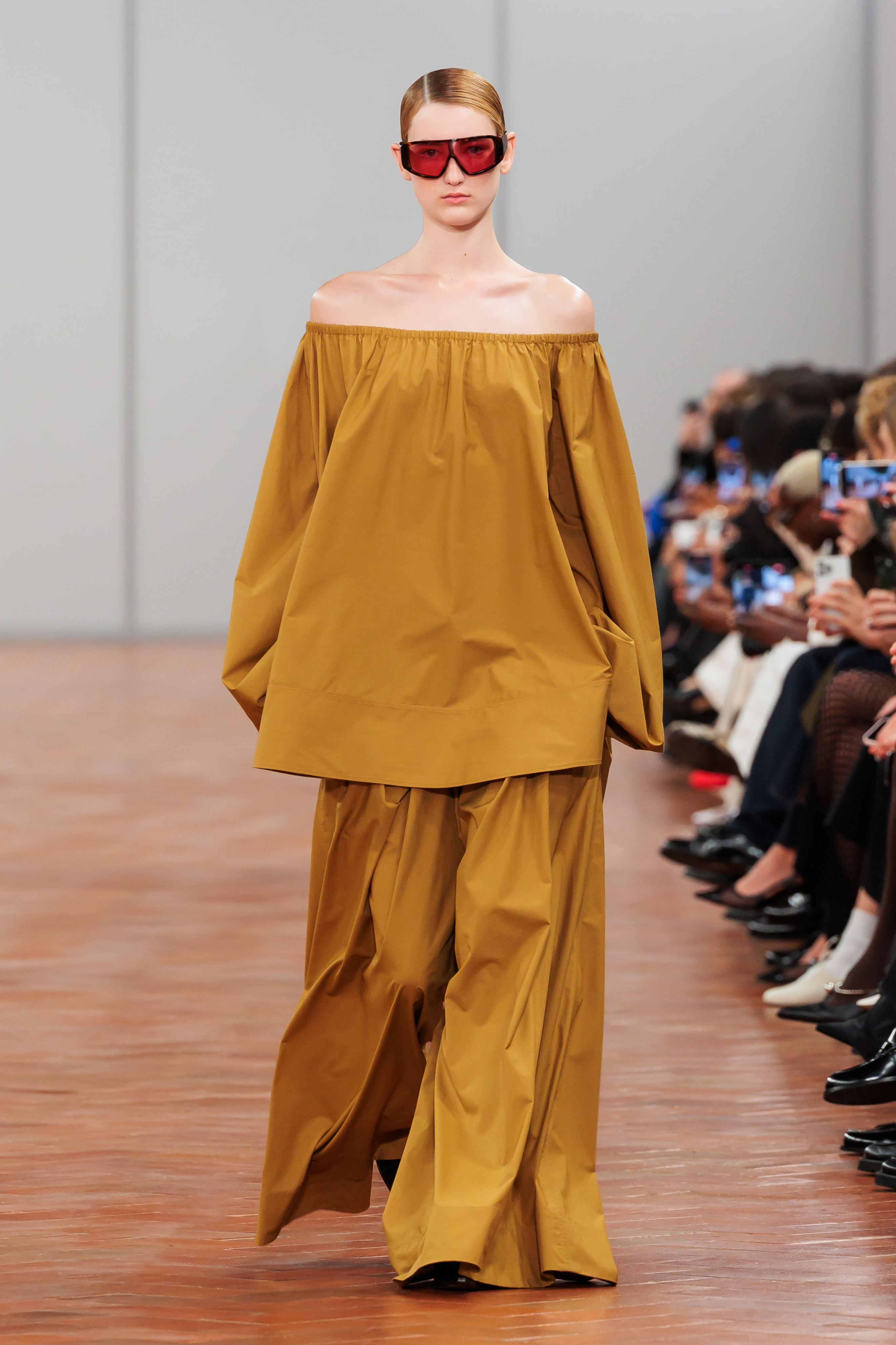 Model on runway wearing an off-shoulder, voluminous top with matching trousers and red sunglasses