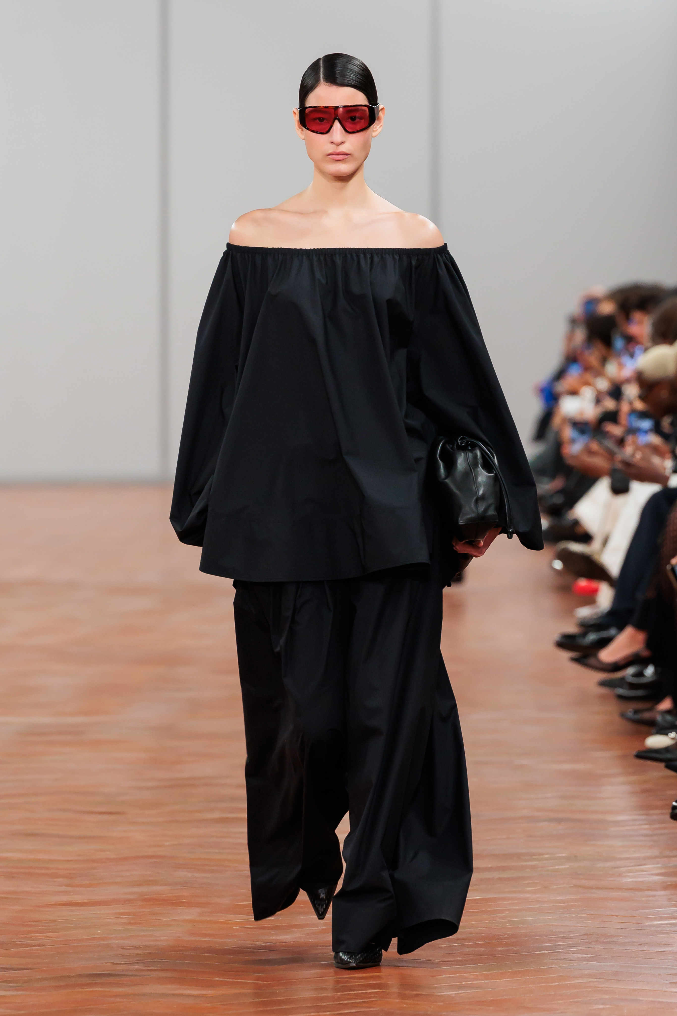 Model on runway wearing an off-shoulder black top, wide-legged pants, carrying a bag, and sporting large sunglasses