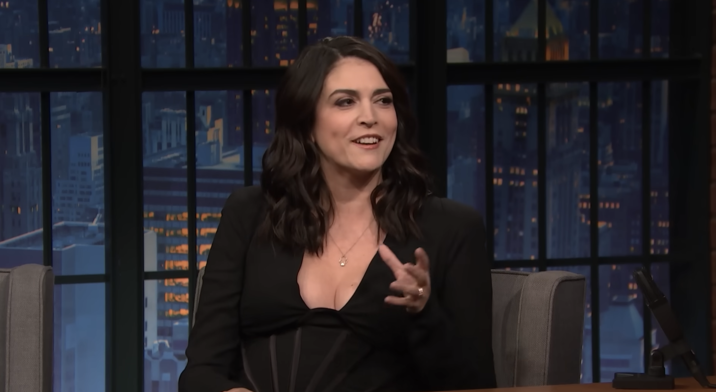 Cecily in a black outfit sitting, gesturing on the talk show