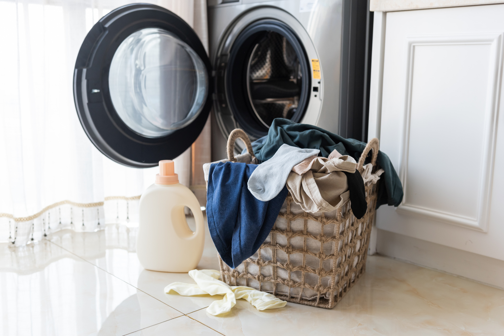 Laundry basket filled with clothes in front of a washing machine, detergent nearby