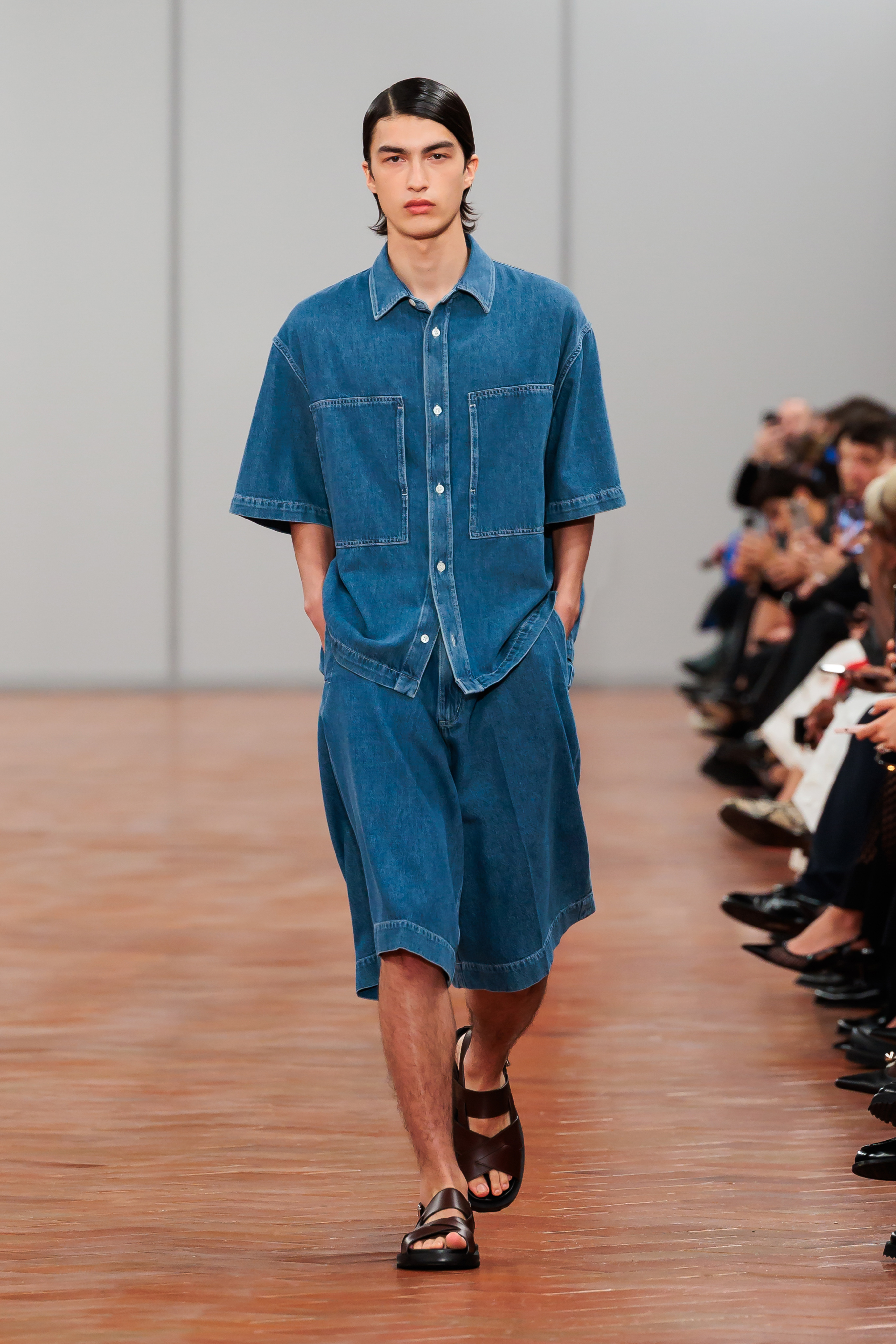 Model on runway wearing oversized denim shirt, matching knee-length shorts, and black sandals. Hands in pockets, stoic expression