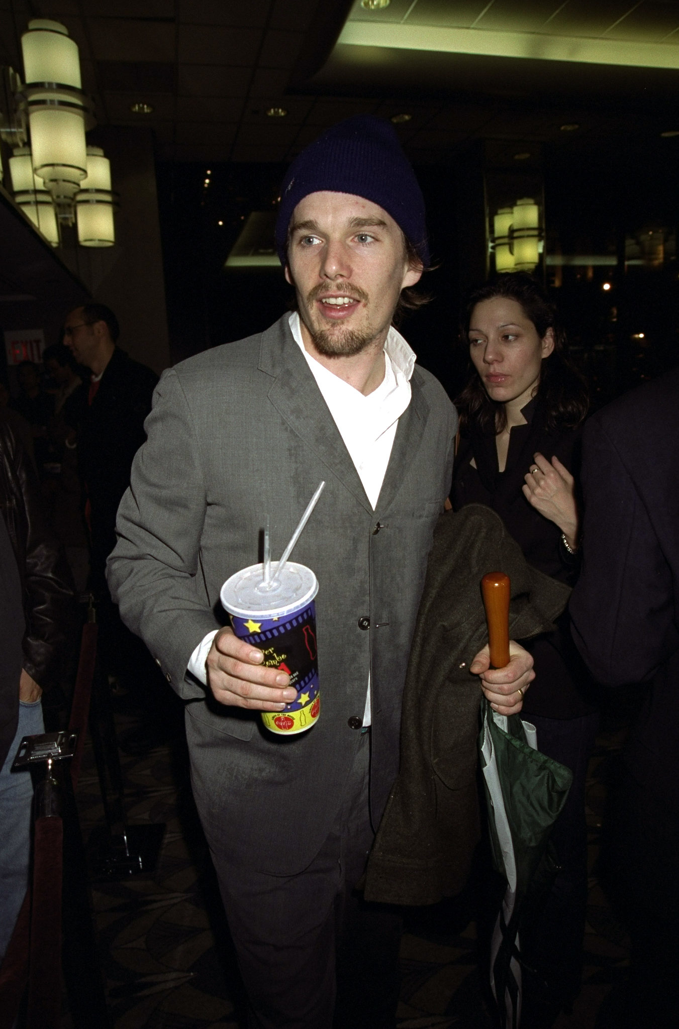 Ethan Hawke in a beanie and casual attire with a beverage and someone beside him at an event