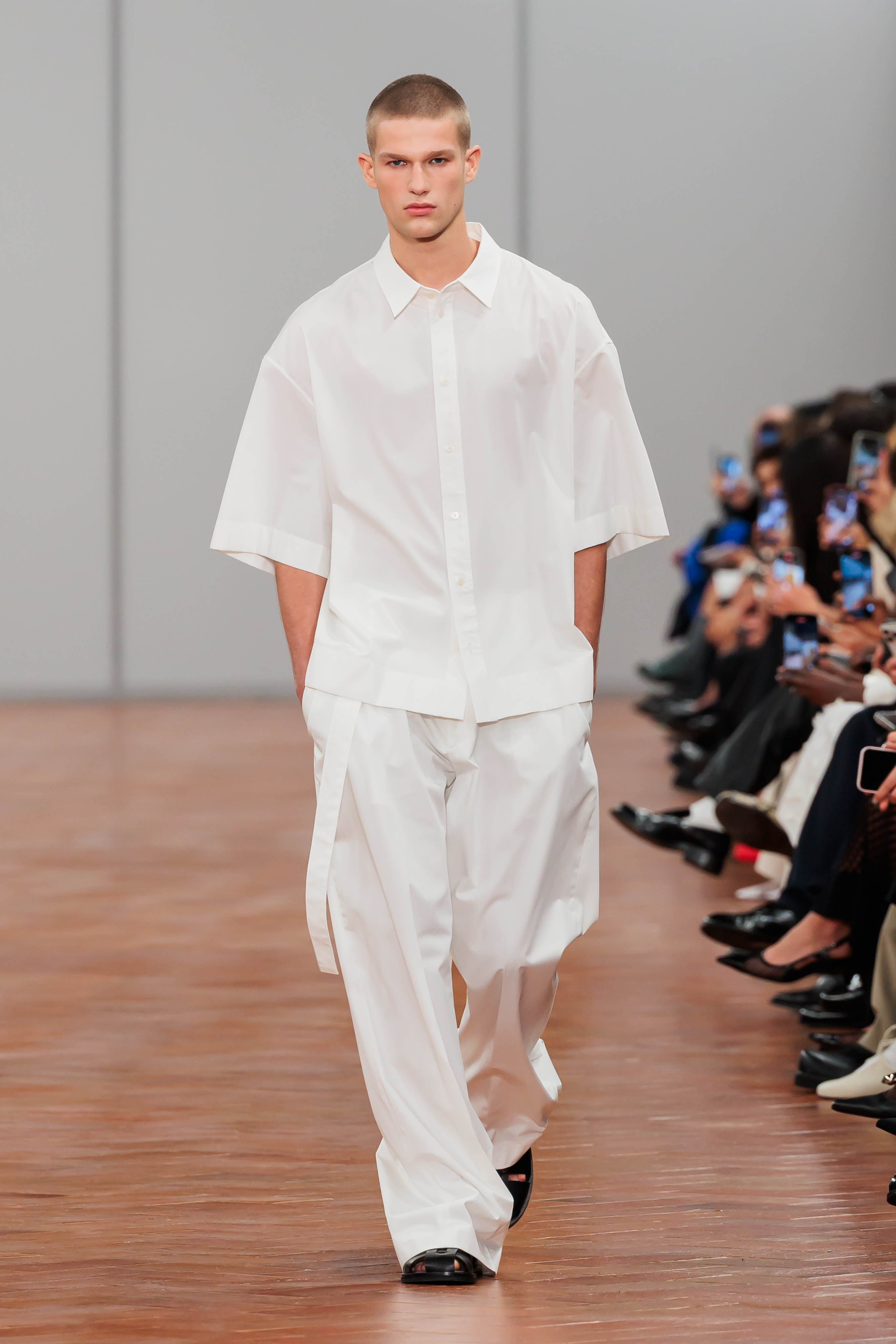 Model in a fashion show wearing an oversized white shirt paired with wide-leg pants and black shoes