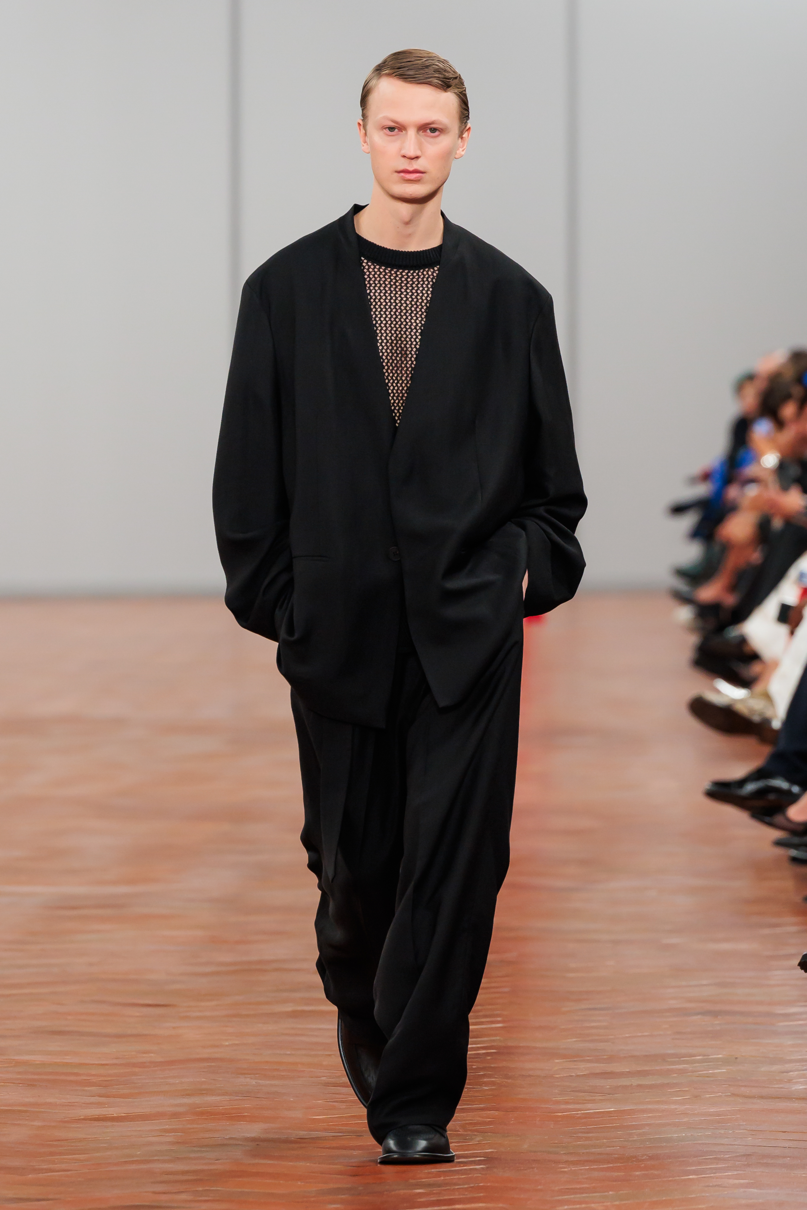 Model on runway in oversized black shirt, sheer mesh top, and wide-leg trousers