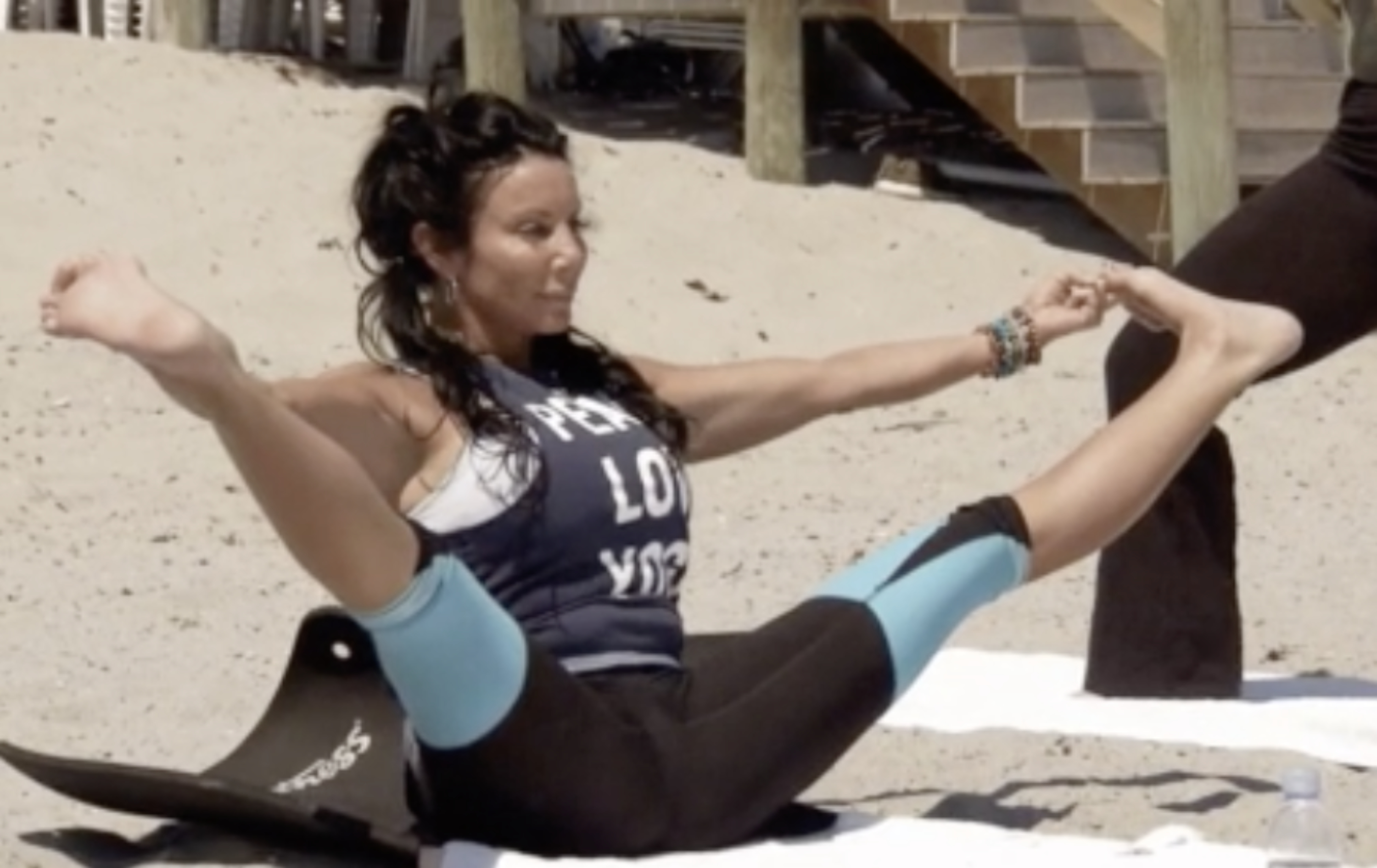 A person performs a yoga pose on the sand, holding their feet, wearing a graphic tank top and two-toned leggings