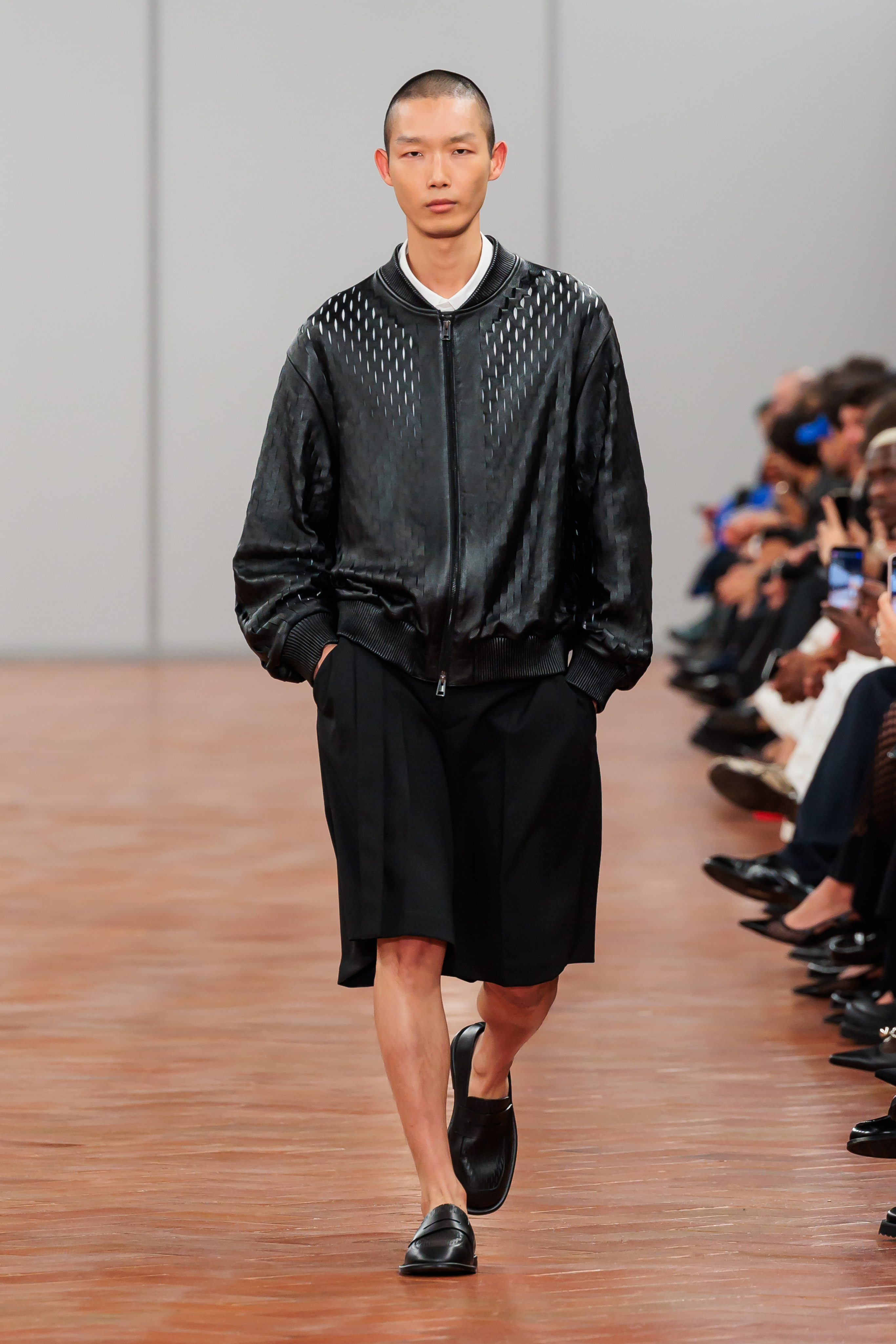 Model walks runway in a perforated leather jacket over a collared shirt and shorts ensemble