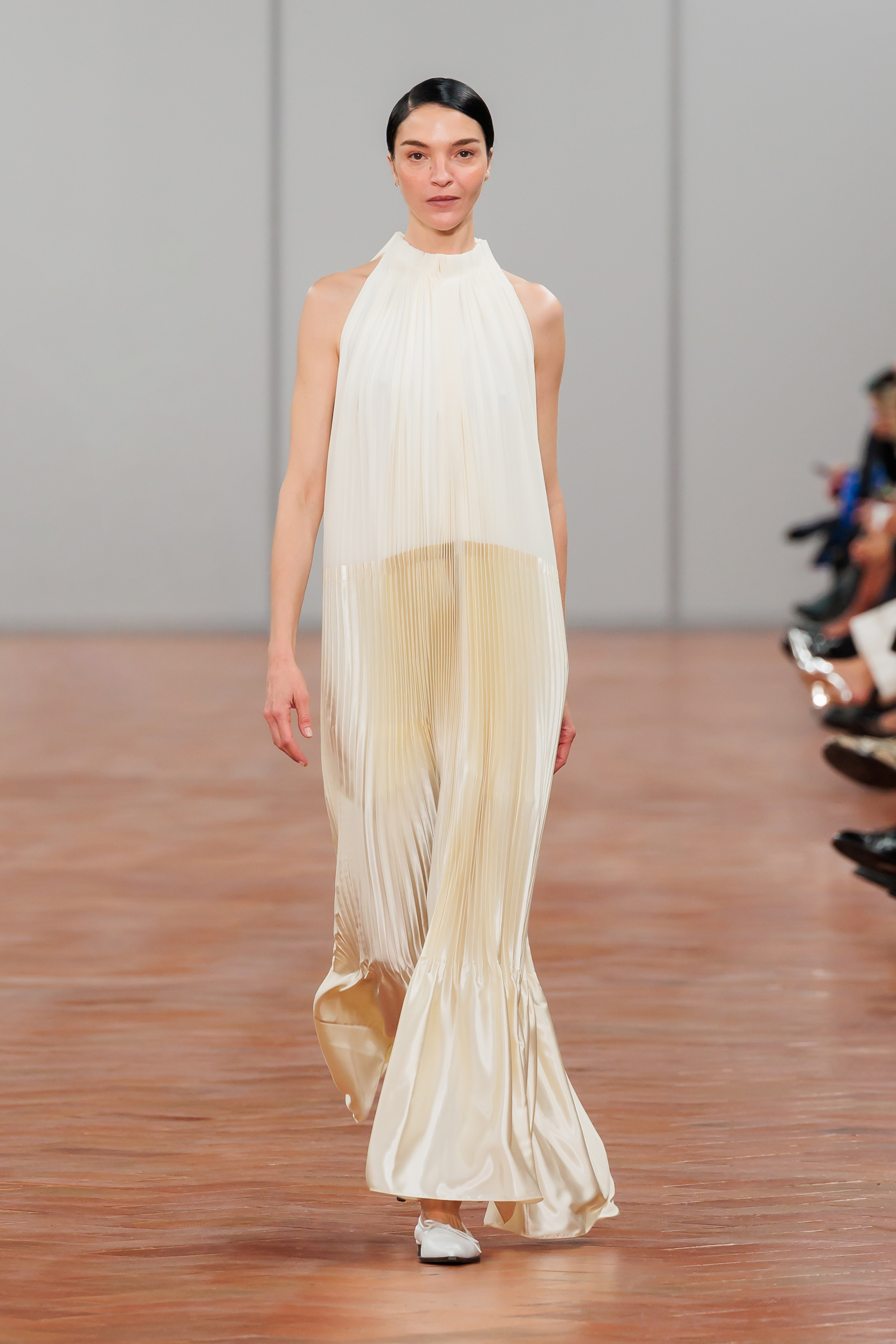 Model on runway wearing a high-neck pleated gown with a draped neckline