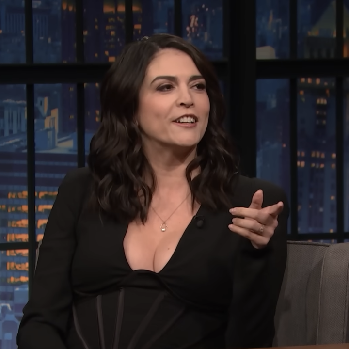 Cecily in a black blazer gestures while sitting on the talk show