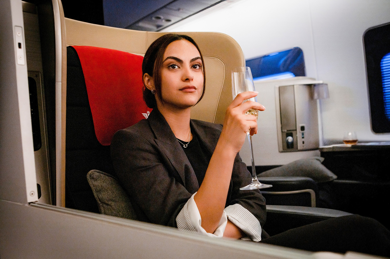 Camila Mendes in business attire holding a champagne glass on an airplane