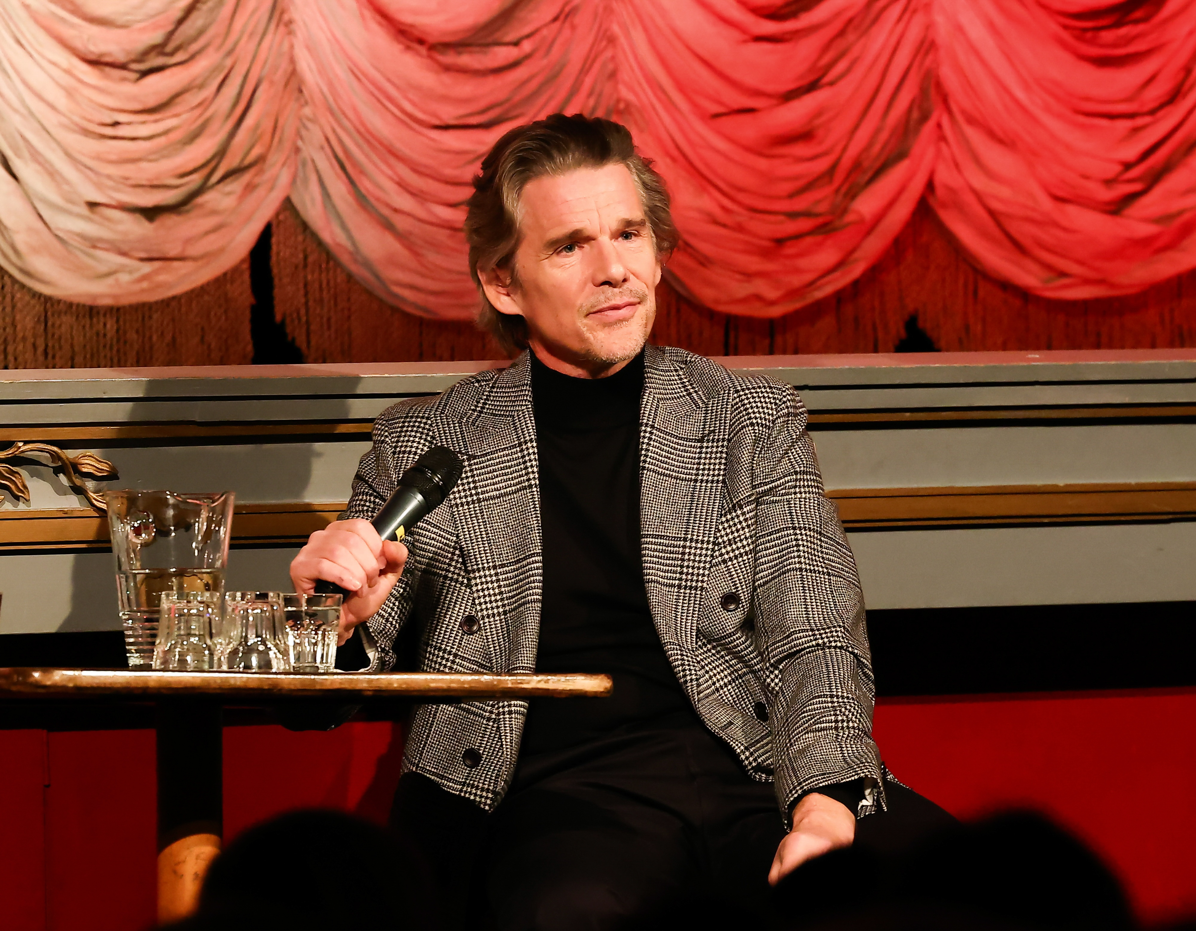 Man in a plaid jacket and black turtleneck seated, holding a microphone, with a red draped background