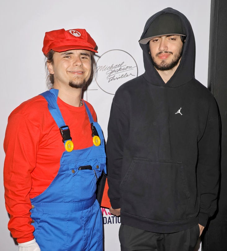 Person on left dressed as Mario, with overalls and cap, person on right in black hoodie and cap, both standing