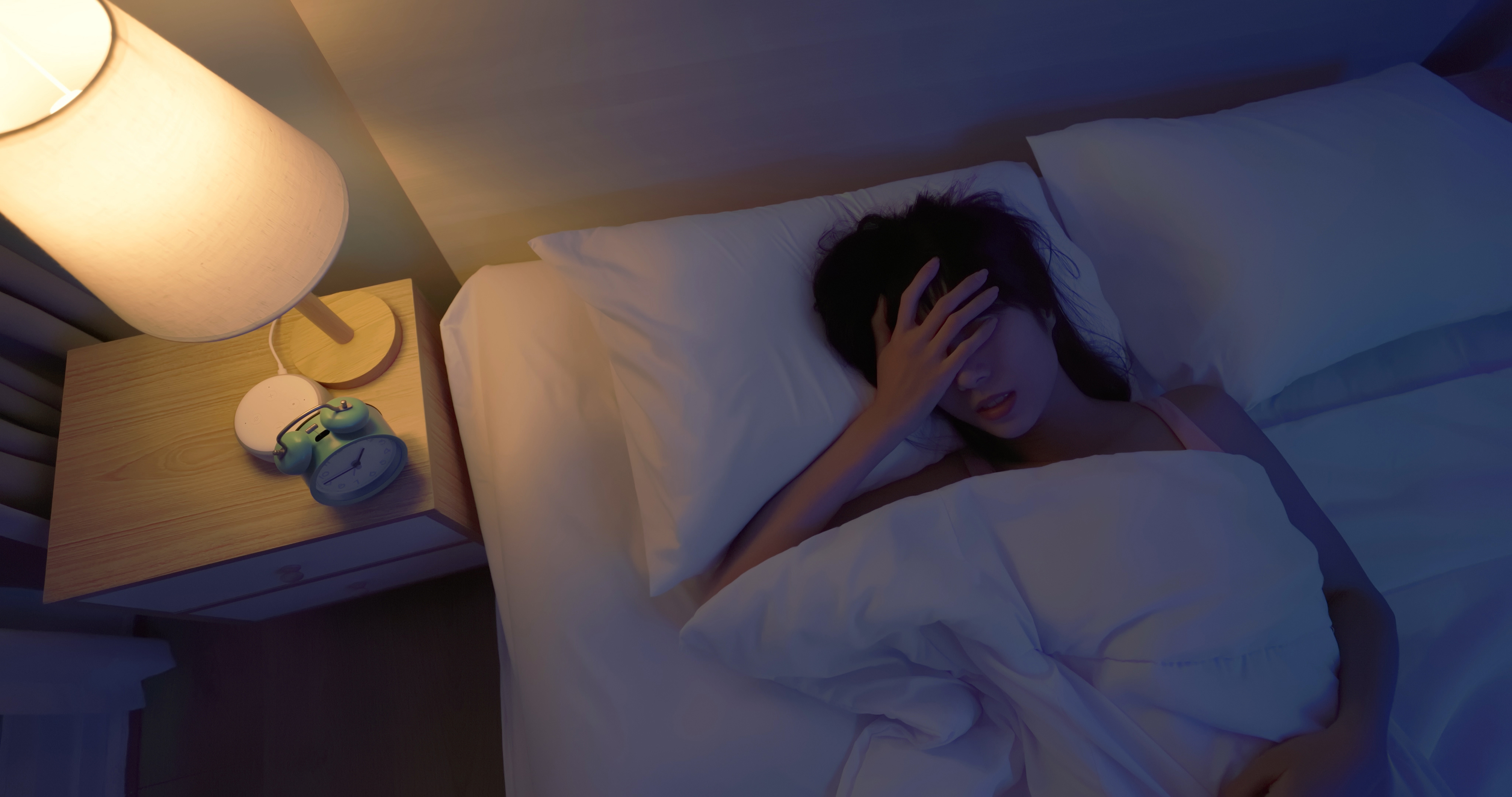 Woman in bed with hand on forehead, alarm clock on nightstand, illustrating sleep troubles for a health article