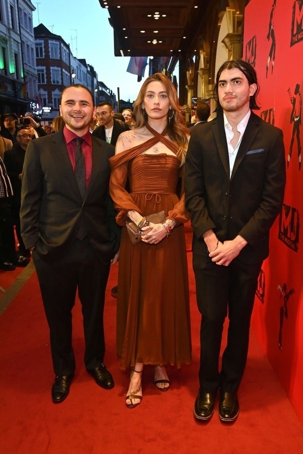 Three people on red carpet, center person in draped brown dress with clasped hands, flanked by men in suits