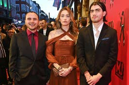 Three people standing together on a red carpet; the center person wears an elegant draped gown, flanked by two in smart suits