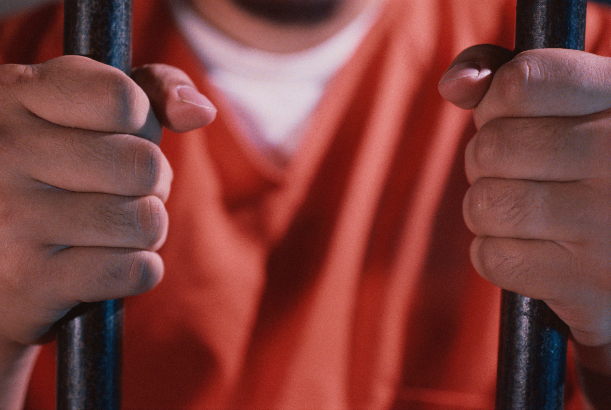 Person holding onto jail bars, focus on hands with an orange prison jumpsuit in the background
