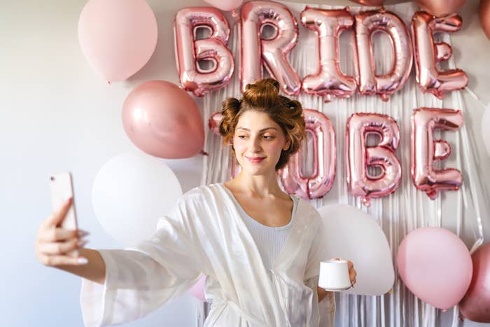 Woman in white robe taking a selfie, with &quot;BRIDE TO BE&quot; balloons in the background