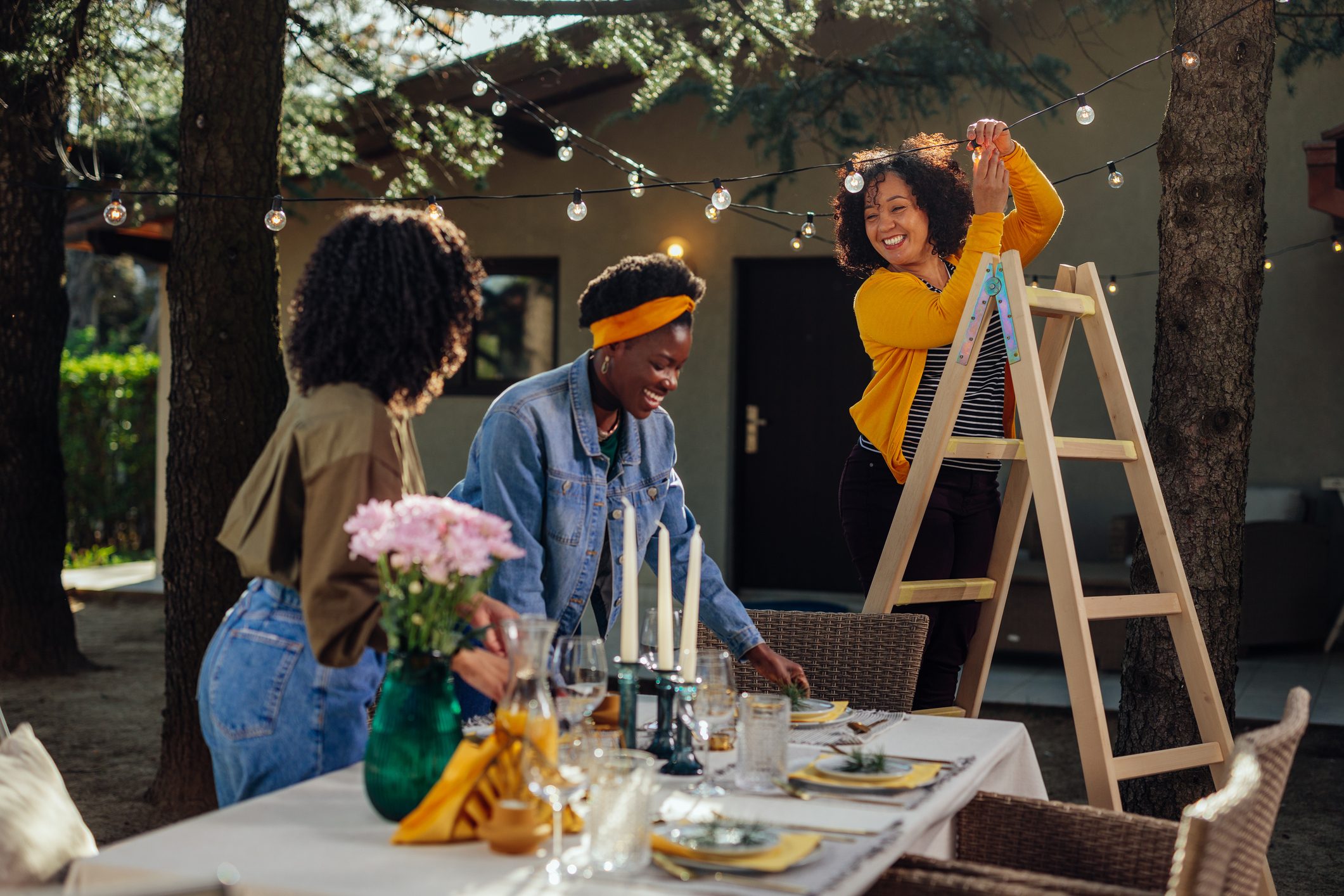Three women setting a table for an outdoor wedding dinner, one arranging flowers, another with a ladder