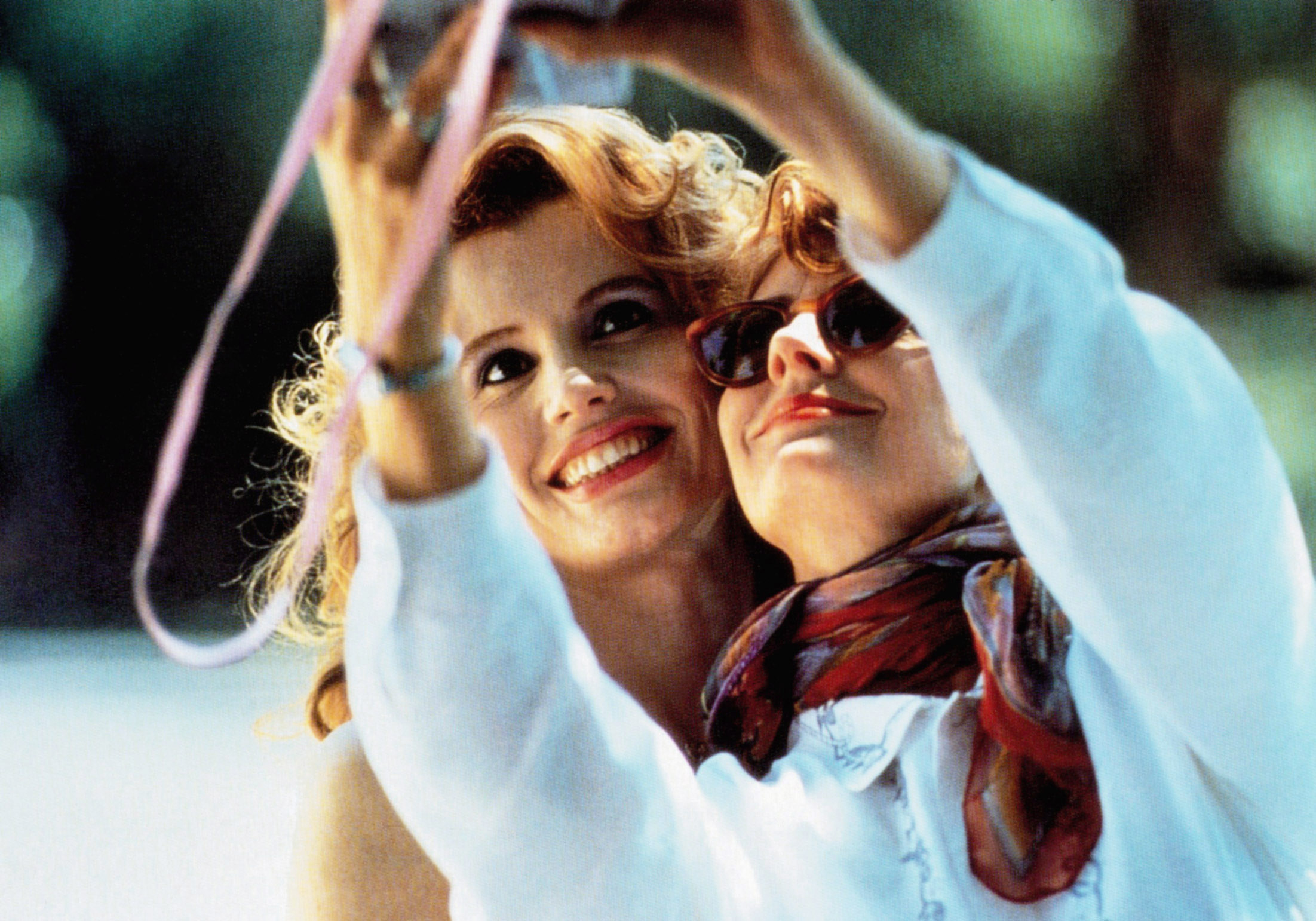 Two women smiling while taking a selfie, one wearing sunglasses and a scarf