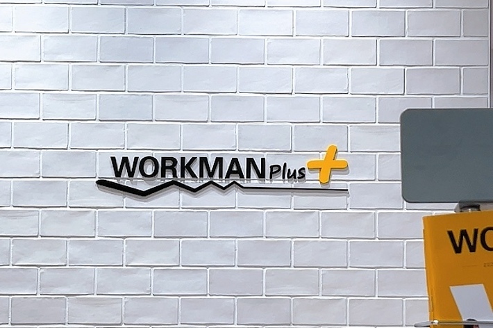Sign saying &lsquo;WORKMAN Plus&rsquo; on a brick-patterned wall, next to a presentation stand
