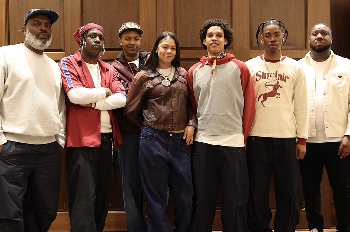 Group of musicians posing together, some wearing casual sweaters and hoodies, with two in front wearing neck scarves
