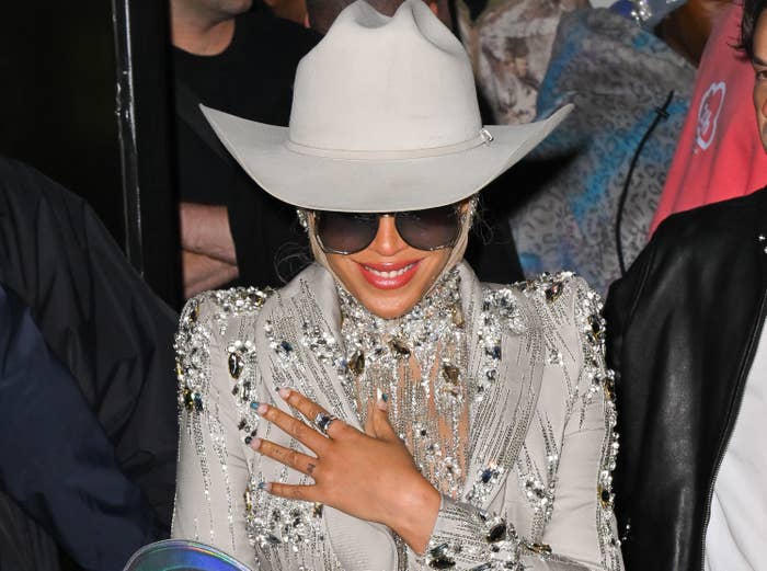 Beyoncé in a wide-brimmed hat and a sparkling jacket with embellishments, smiling behind sunglasses
