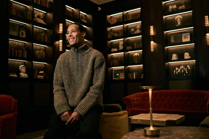 Man in knit sweater smiling, seated in well-lit room with bookshelves and sculptures