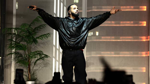 Drake onstage, arms spread, in a black leather jacket and pants, performing with a tropical plant on the side