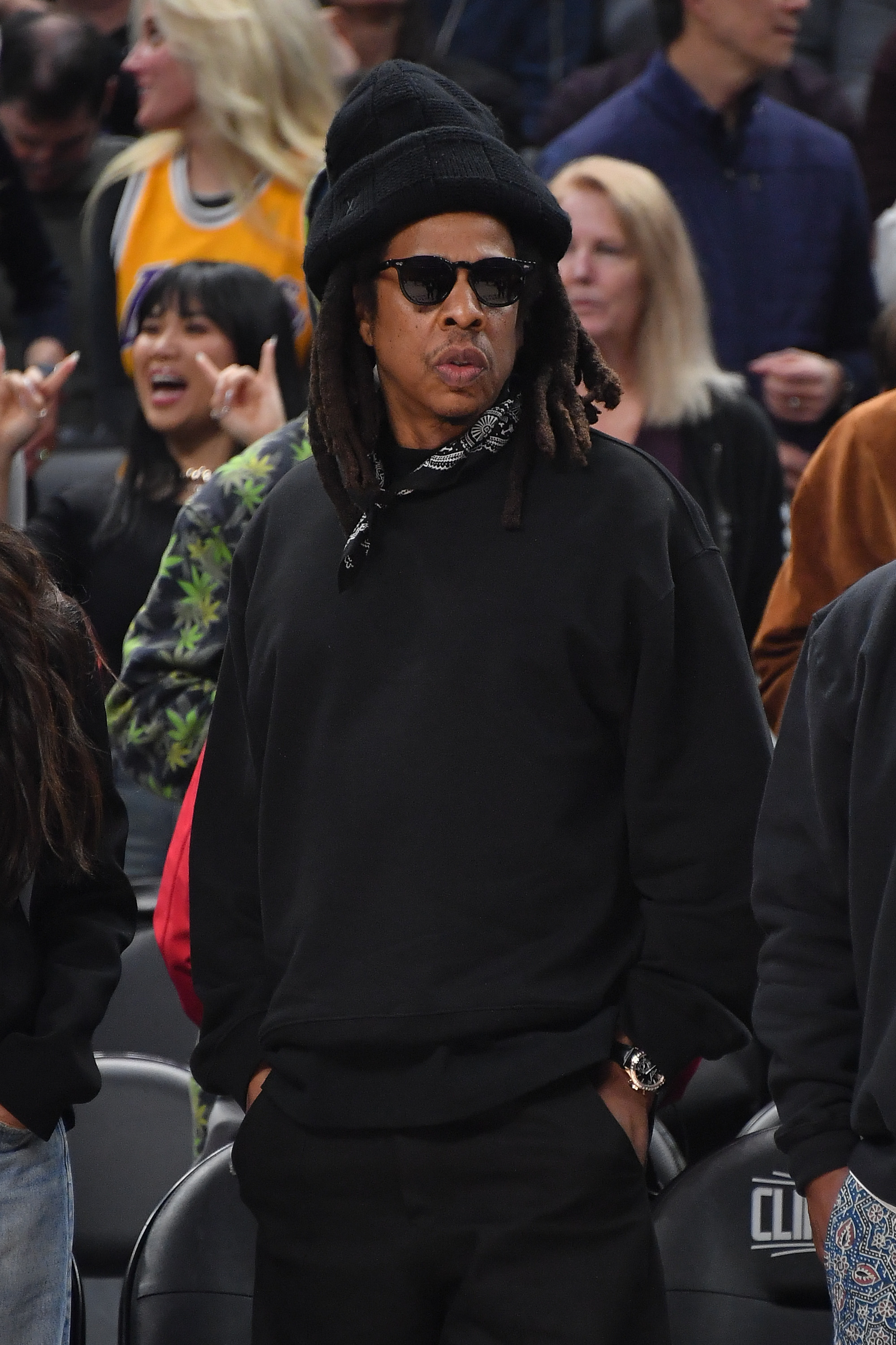 Jay-Z in dark attire, sunglasses, and hat at a sports event with spectators in the background