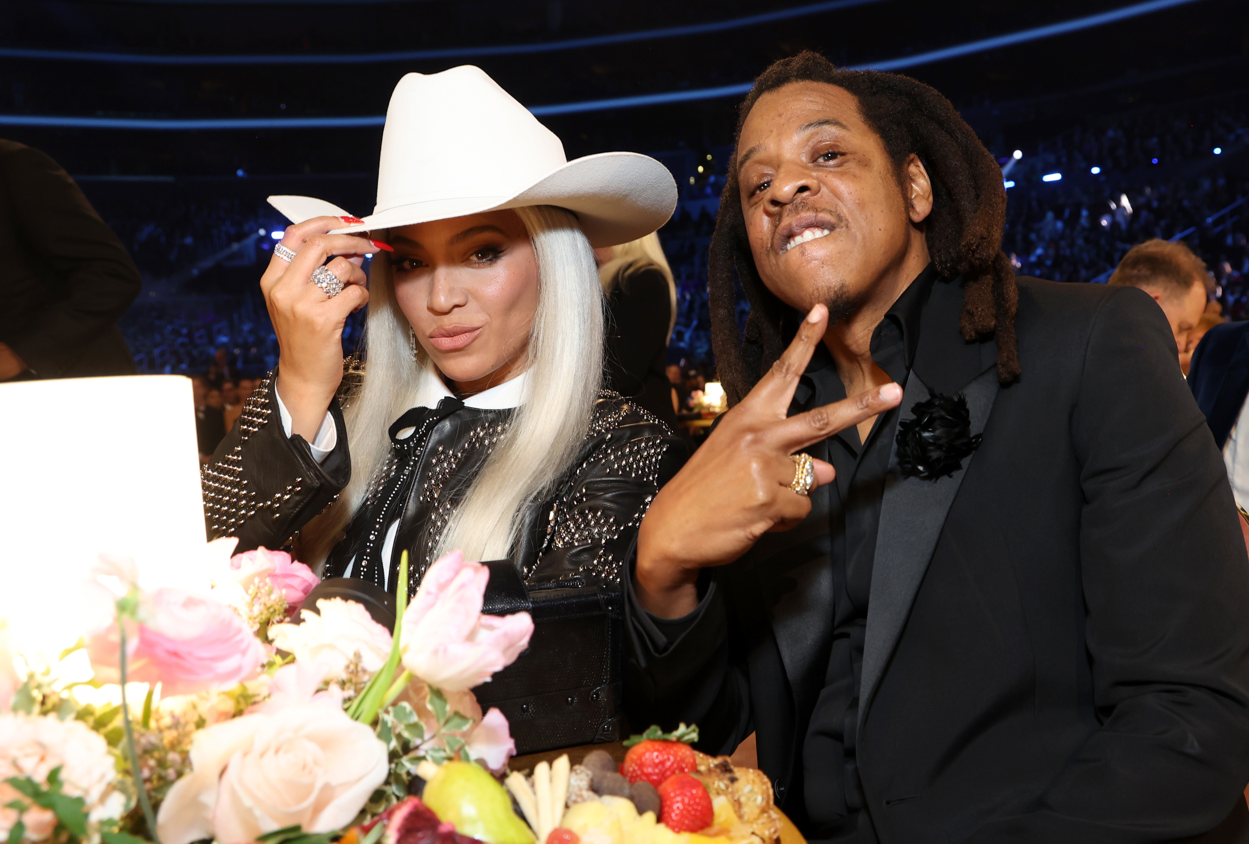 Beyoncé and Jay-Z seated at a table with floral arrangement, she wearing a large cowboy hat and he making a peace sign