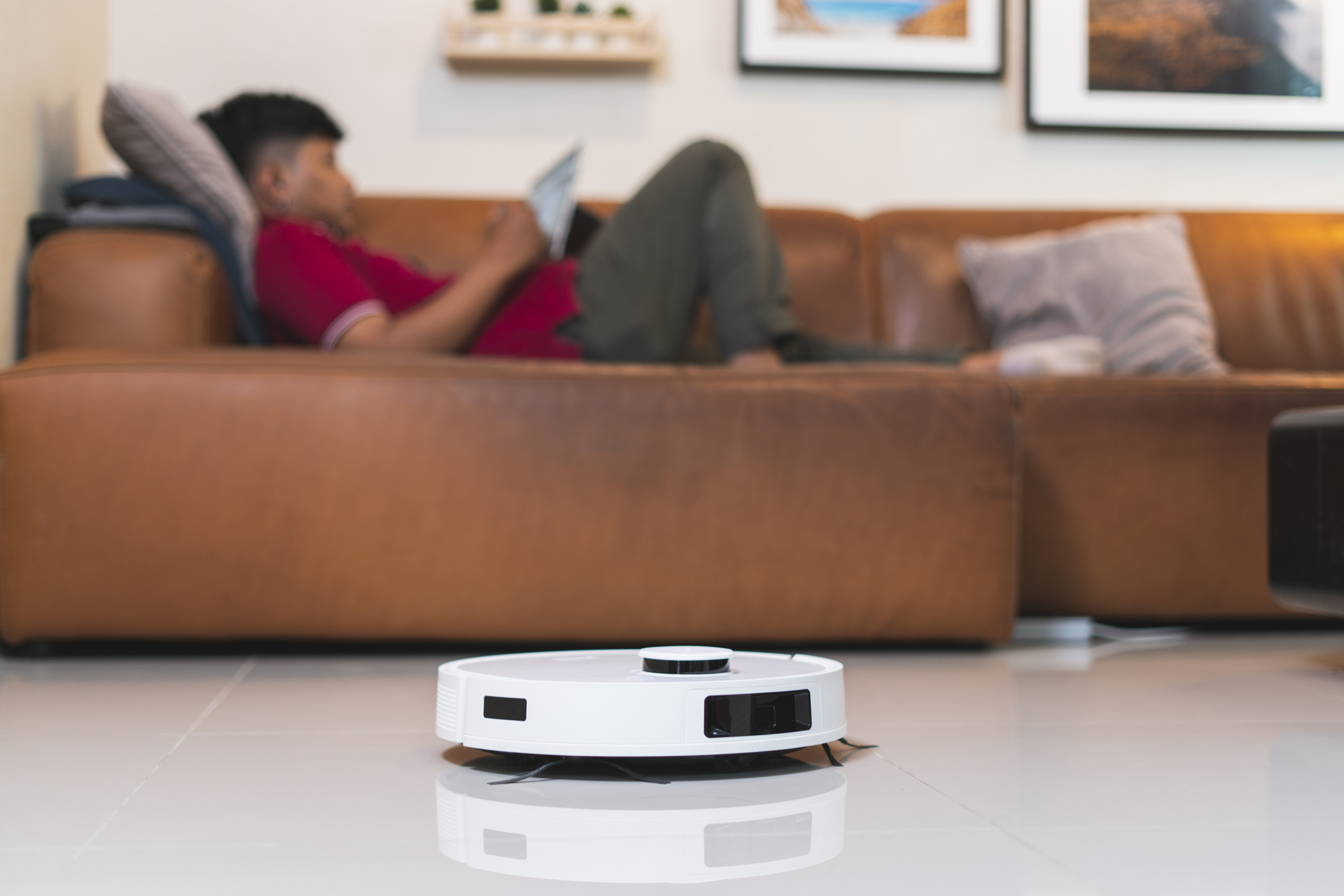 Person relaxing on a couch using a tablet with a robotic vacuum cleaner in the foreground