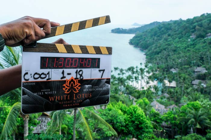 A pair of hands holding a clapperboard for &quot;The White Lotus&quot; in front of a lush coastal view