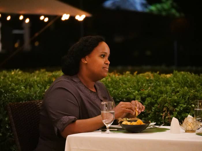 Natasha Rothwell at a table with a meal, looking off to the side, with string lights and greenery in the background