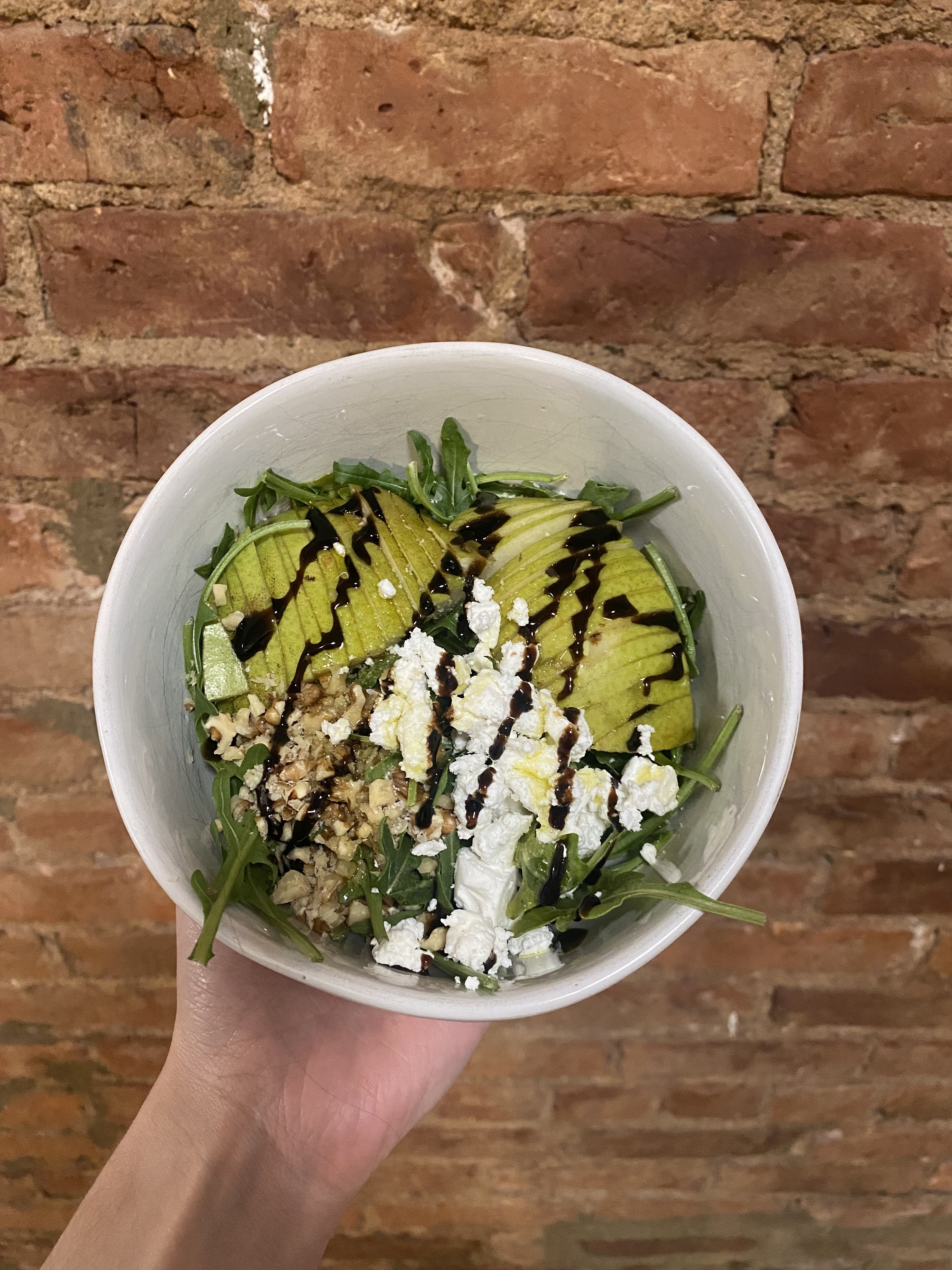 Person holding a bowl with salad, topped with avocado, cheese, and a dark dressing drizzle. Brick wall in background