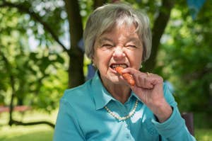 Elderly woman grimacing while biting into a whole carrot, outdoors during daytime