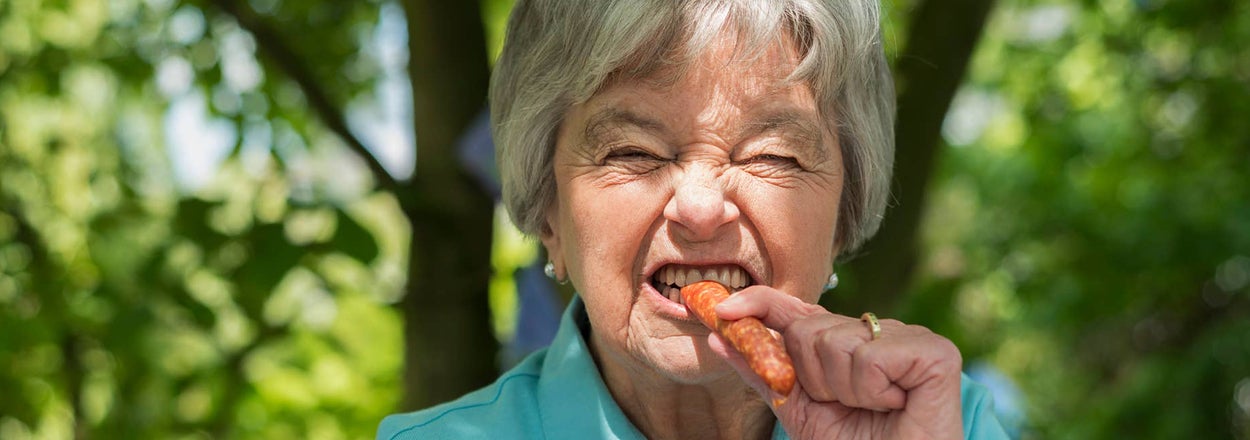 Elderly woman grimacing while biting into a whole carrot, outdoors during daytime