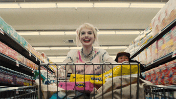 gif of harley quinn with a big smile riding in a grocery cart in a supermarket aisle in movie birds of prey