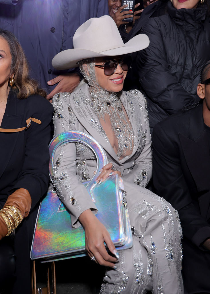 Beyoncé in a beaded pantsuit with a matching cowboy hat, smiling, holding a metallic purse