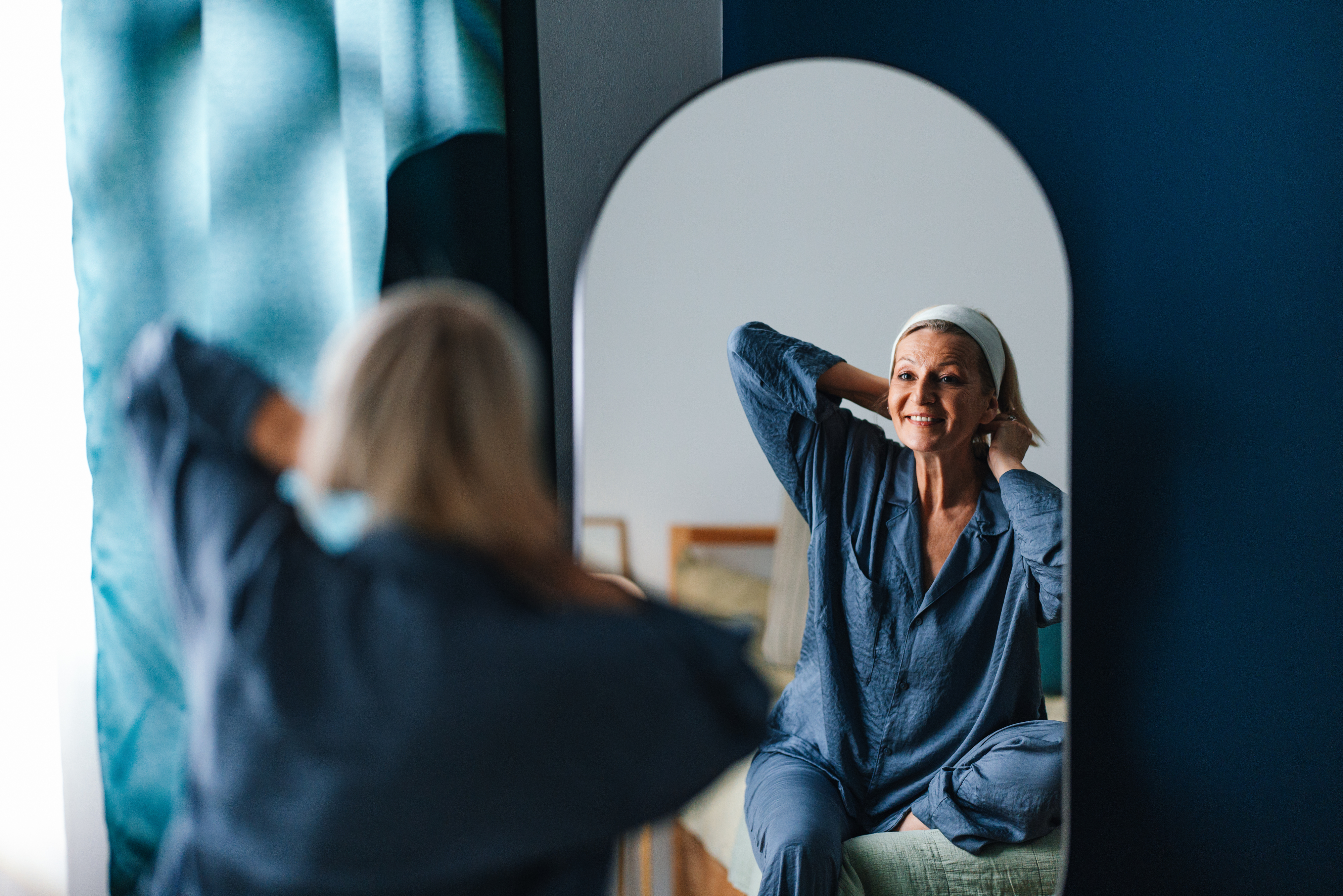 Woman in pajamas smiles at her reflection in a bedroom mirror