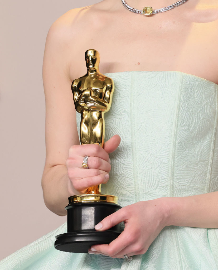 Person holding an Oscar award, wearing a light-textured gown with a diamond ring