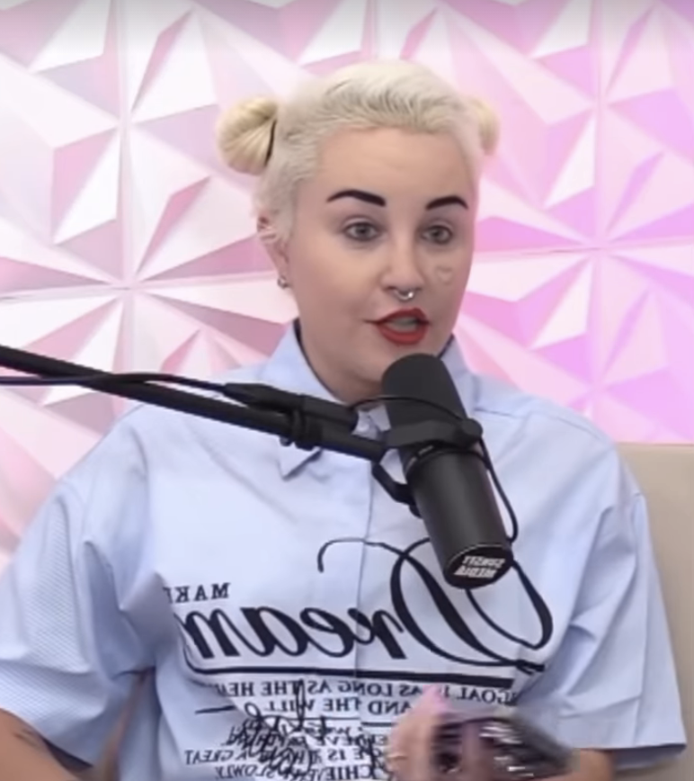 Amanda with styled blonde hair, in a buttoned shirt, sitting in front of a microphone with a geometric background