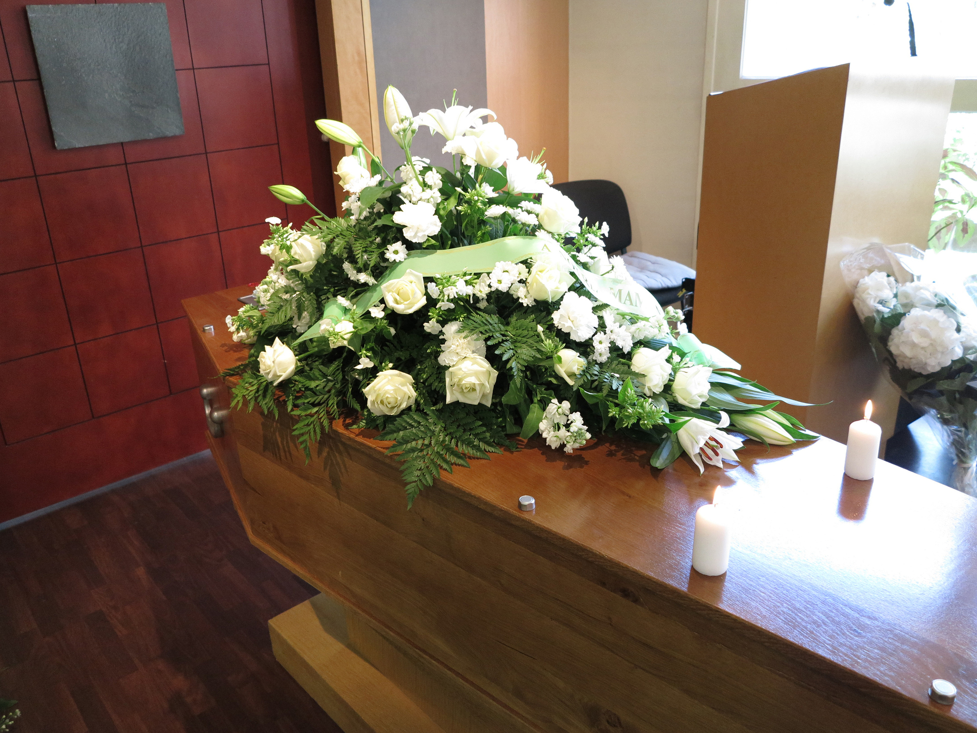 A closed wooden casket with white floral arrangements on top, flanked by lit candles, in a room