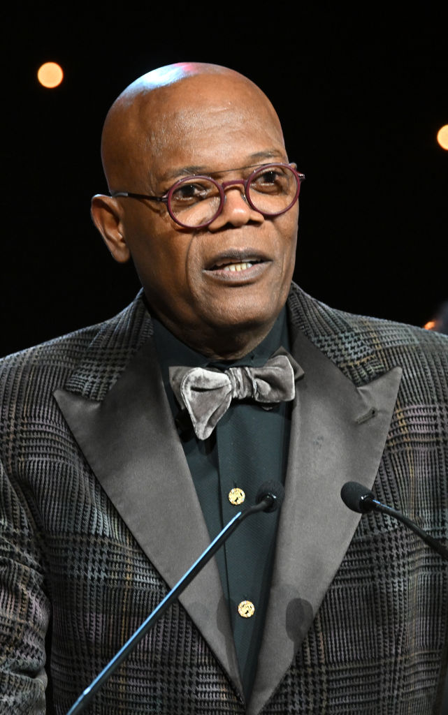 Samuel L. Jackson at an event wearing a patterned suit and velvet bow tie, speaking at a podium