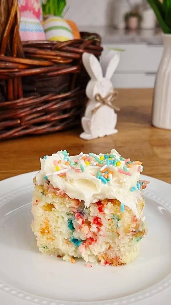 A slice of funfetti cake with white frosting and sprinkles on a plate, a basket and bunny decor in the background