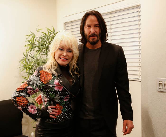 Dolly Parton and Keanu Reeves posing together, Dolly in a floral embellished black outfit, Keanu in a black suit