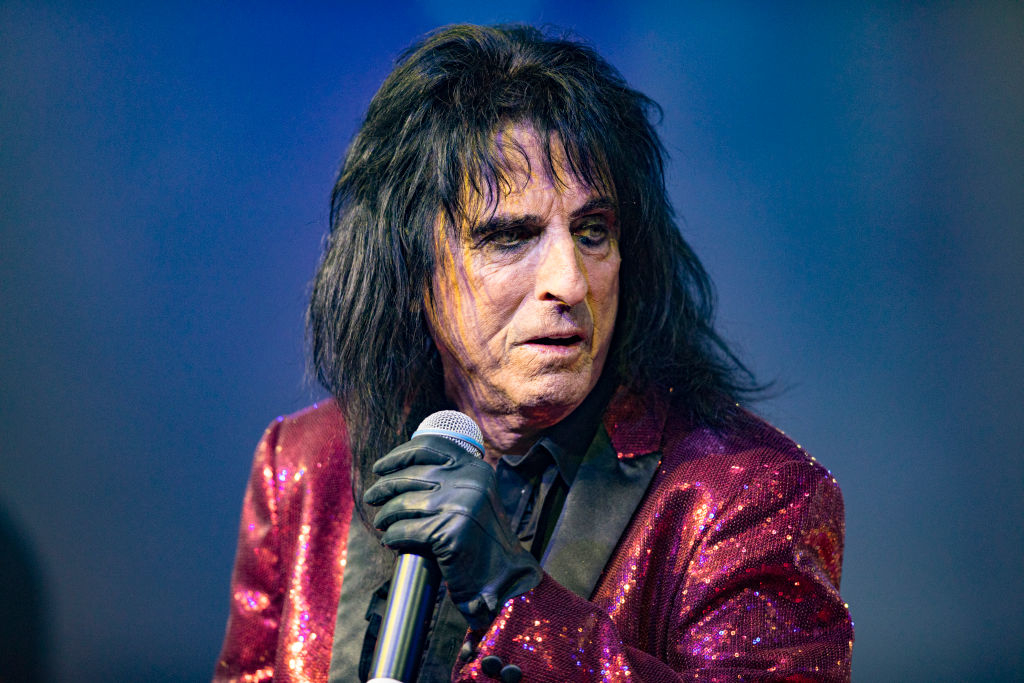 Person on stage with mic, wearing a sparkly jacket, black shirt, and heavy eye makeup